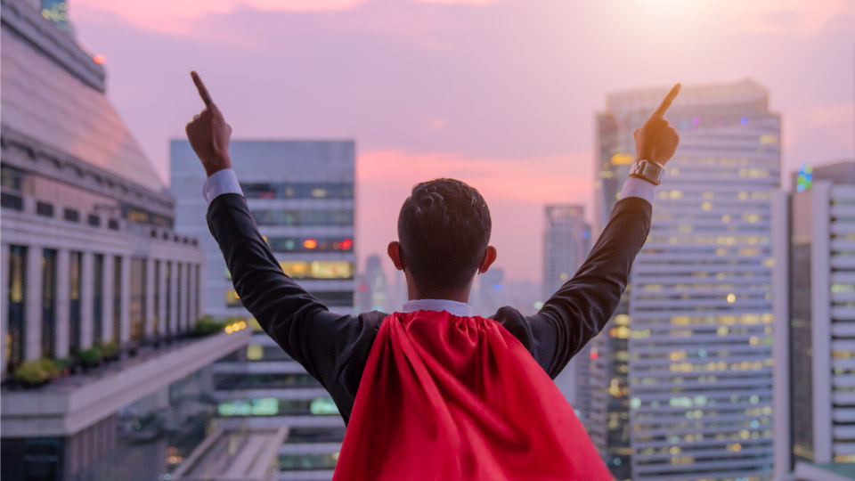 man with red cape looking over city skyline with his hands raised