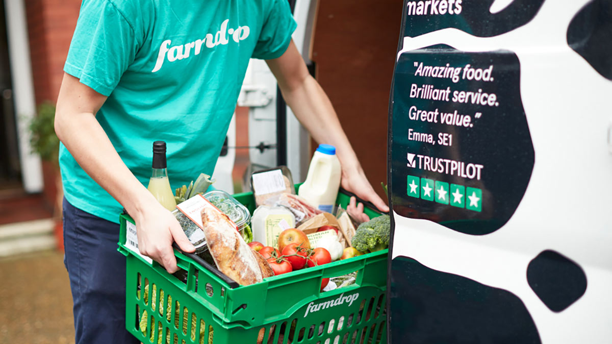 A person carrying a green shopping basket filled with produce and groceries.