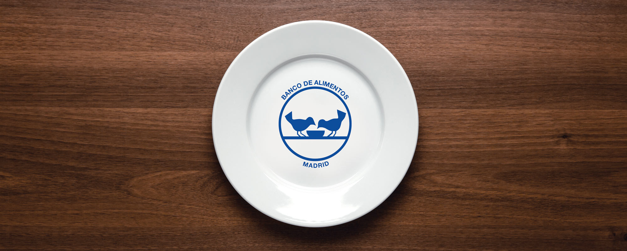 White plate on table with blue design on it of two birds eating out of a bowl with the text 'Banco De Alimentos Madrid'
