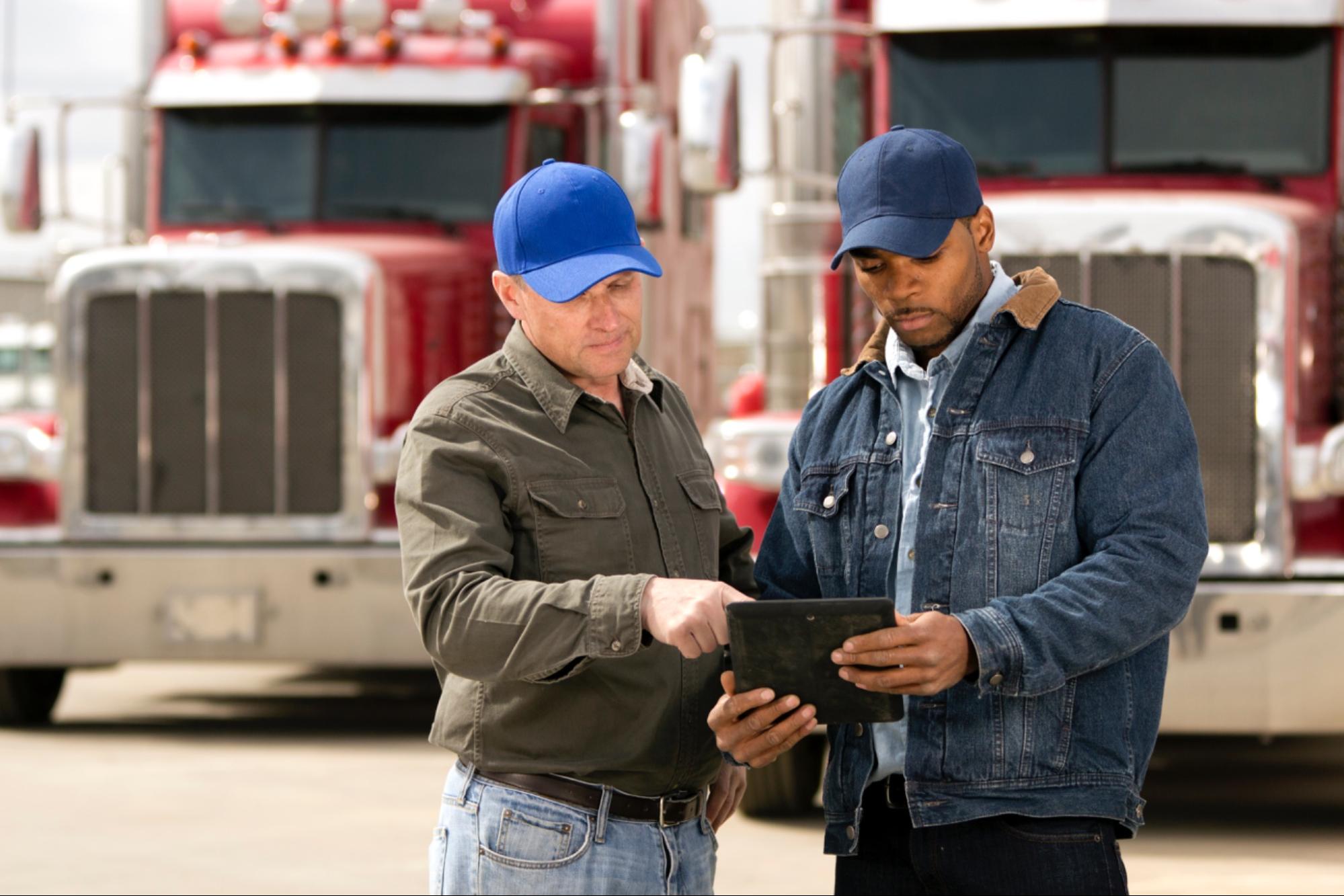 Deux chauffeurs routiers consultant une tablette devant des camions rouges.