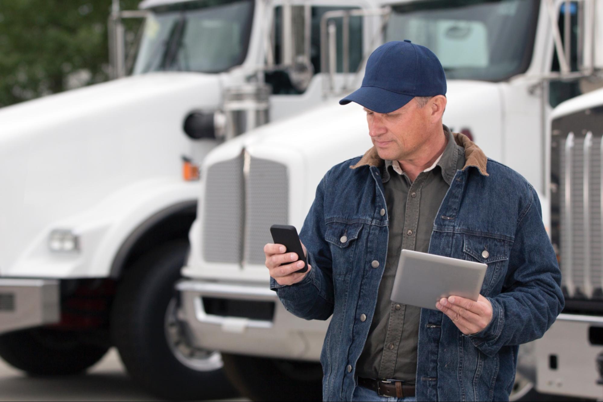 Chauffeur routier utilisant un smartphone et une tablette devant des camions blancs.