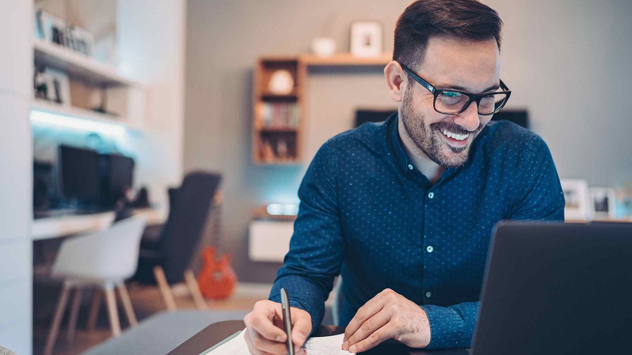 Foto de hombre joven con lente, vestido de azul, viendo sonriente la pantalla de su computadora portatil