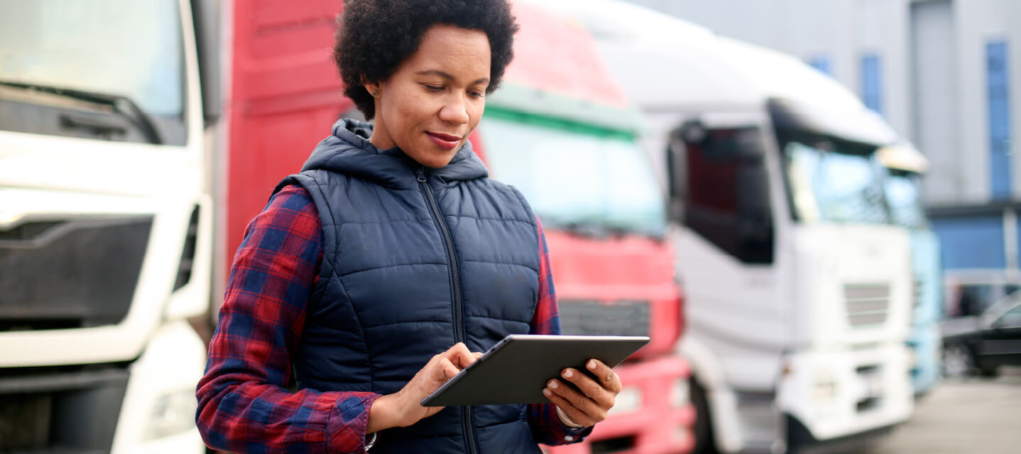 Person in their 30s standing in front of a row of parked semi-trucks outside a distribution warehouse, holding a digital tablet.