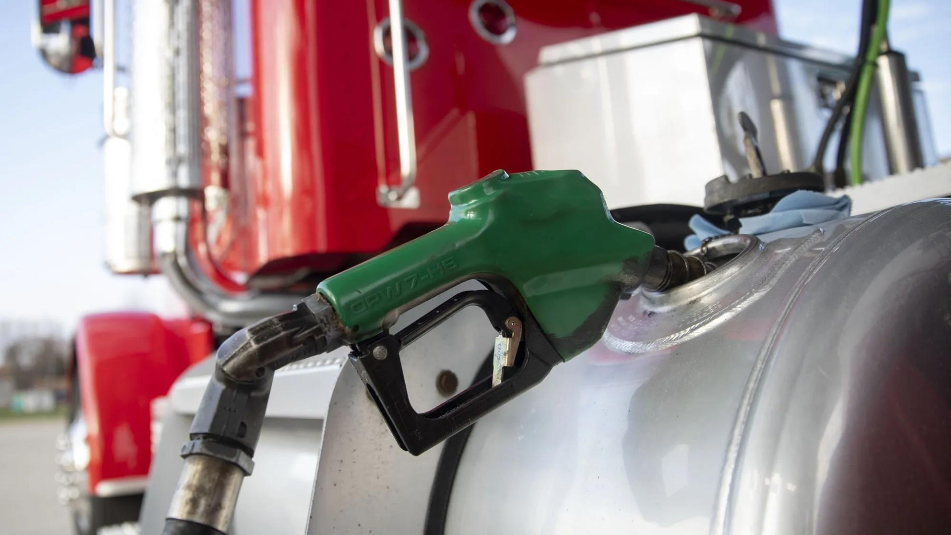 A green fuel pump nozzle inserted into a silver tanker truck at a gas station with red industrial equipment blurred in the background