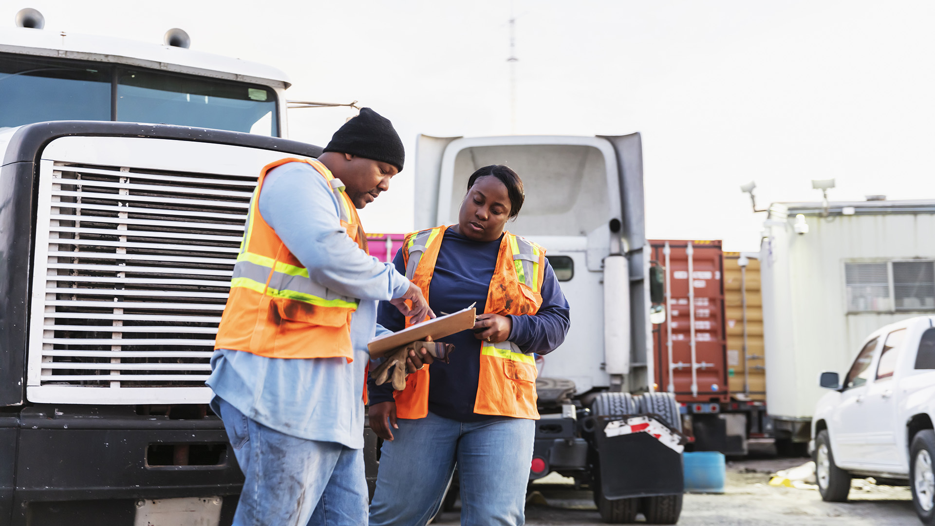 Two fleet workers in safety vests reviewing notes on a clipboard beside a truck