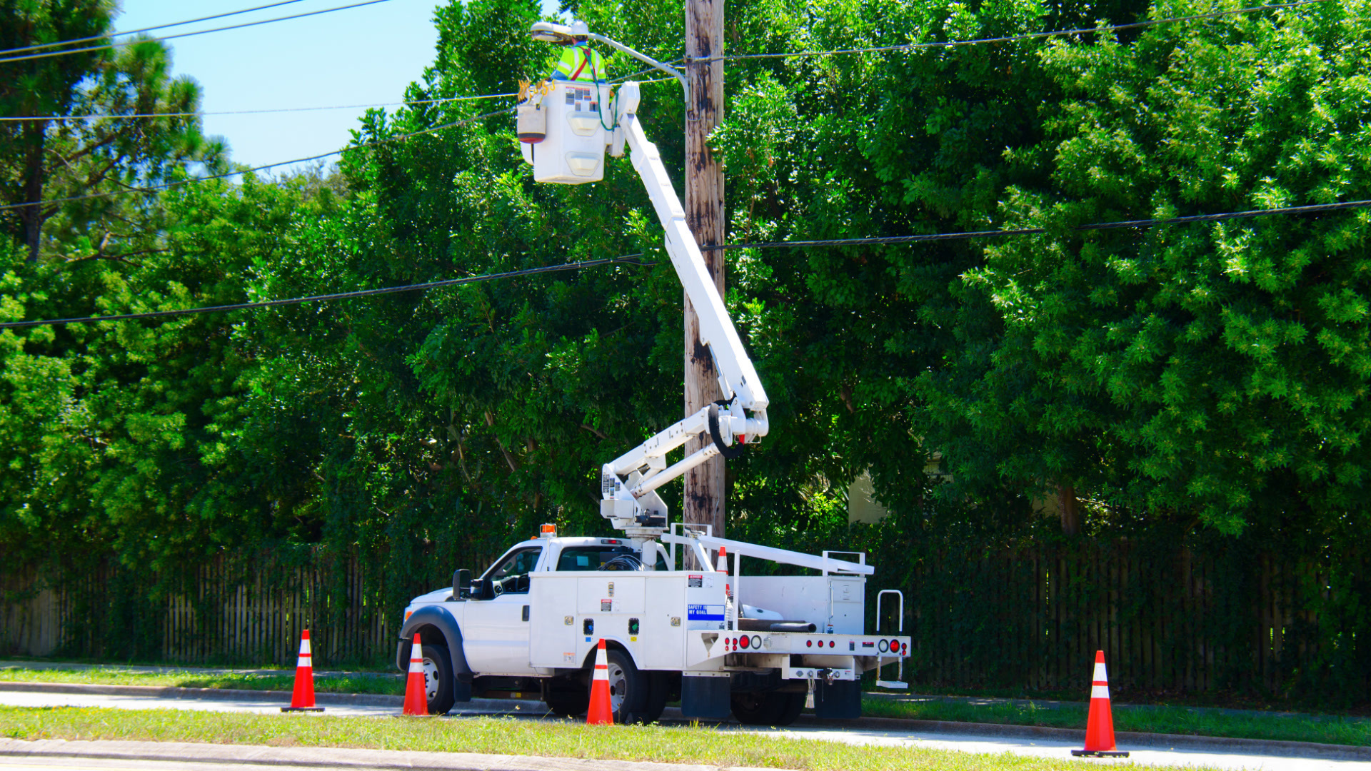 Utility bucket truck parked at the roadside with a lineworker in the elevated bucket servicing overhead power lines.
