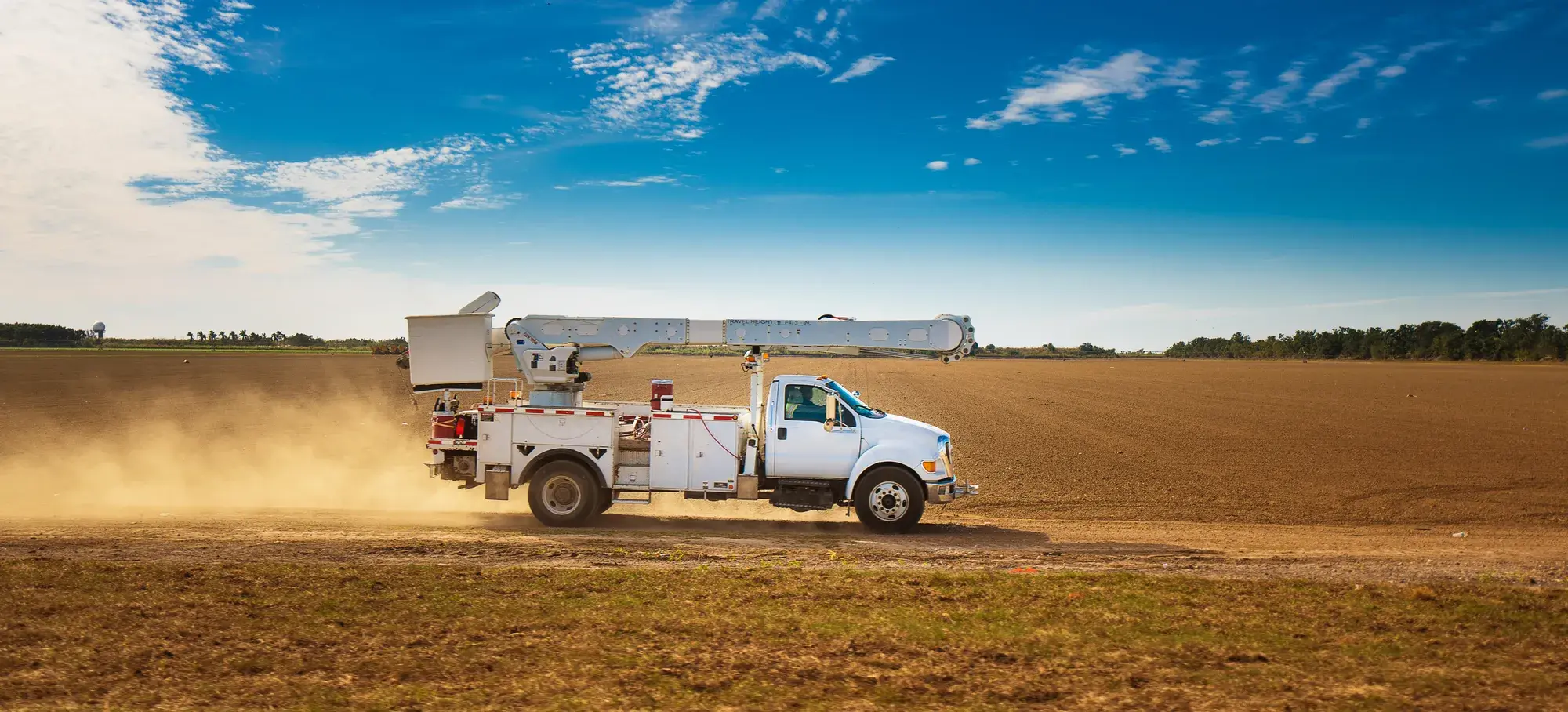 White utility bucket truck driving across a dusty field under a blue sky