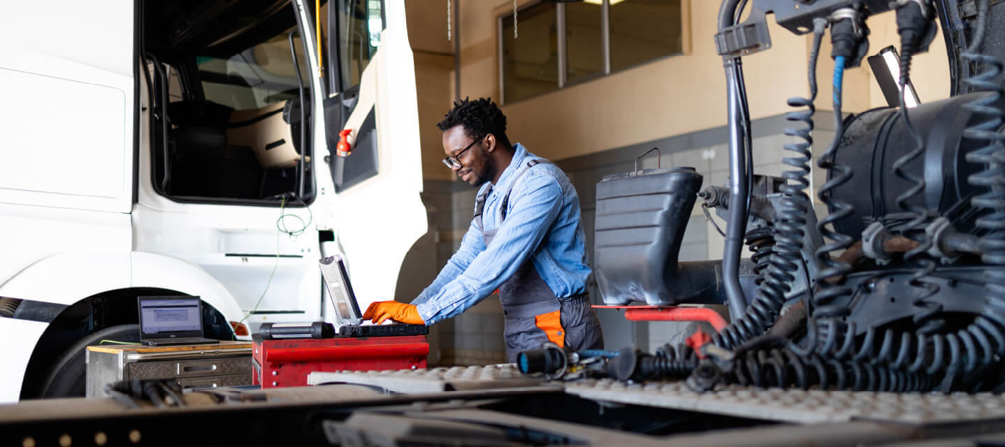 A man standing next to a fleet truck inside a garage, working on a computer. 