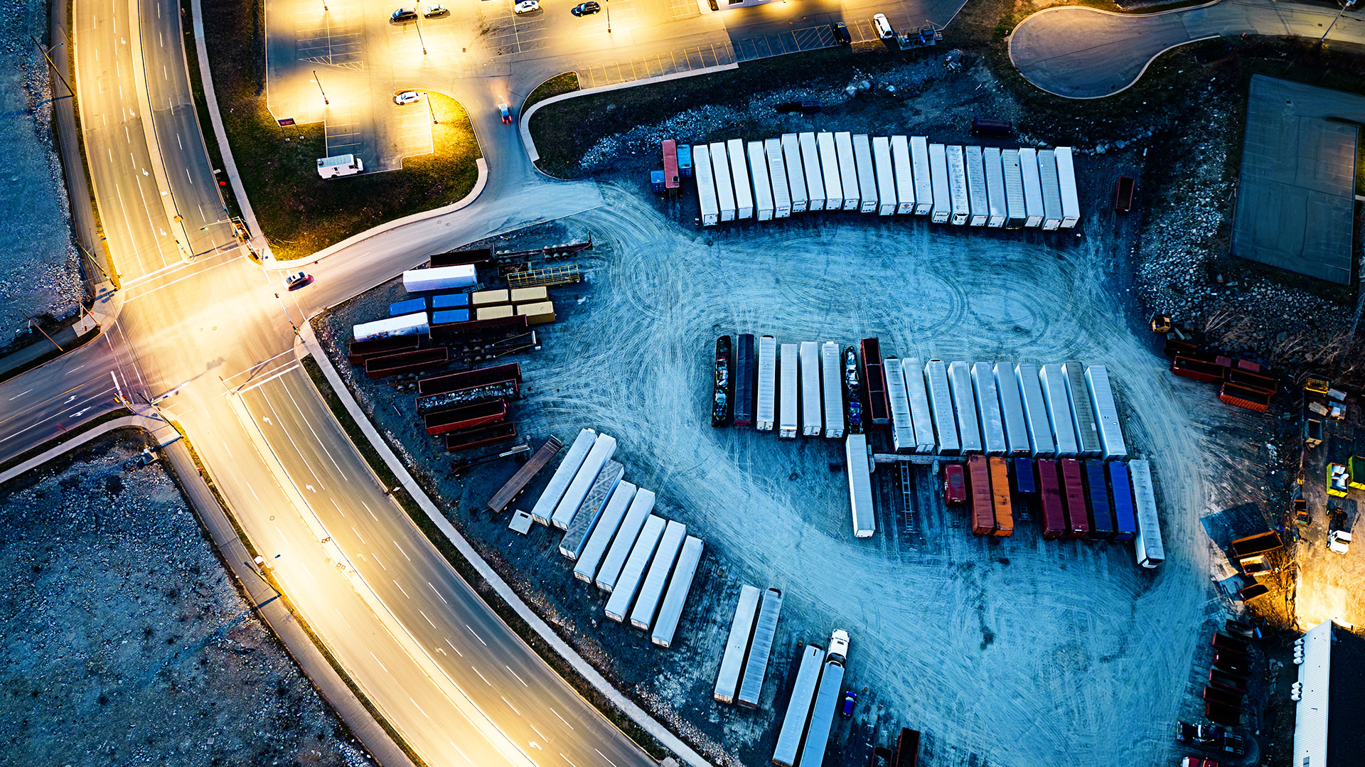 Aerial view of a logistics yard at dusk with dozens of parked semi-trucks and shipping containers arranged in rows on pavement, adjacent to an illuminated road