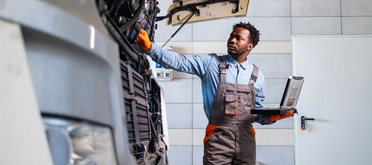  A man in maintenance gear and orange gloves working on a fleet vehicle and holding a laptop. 