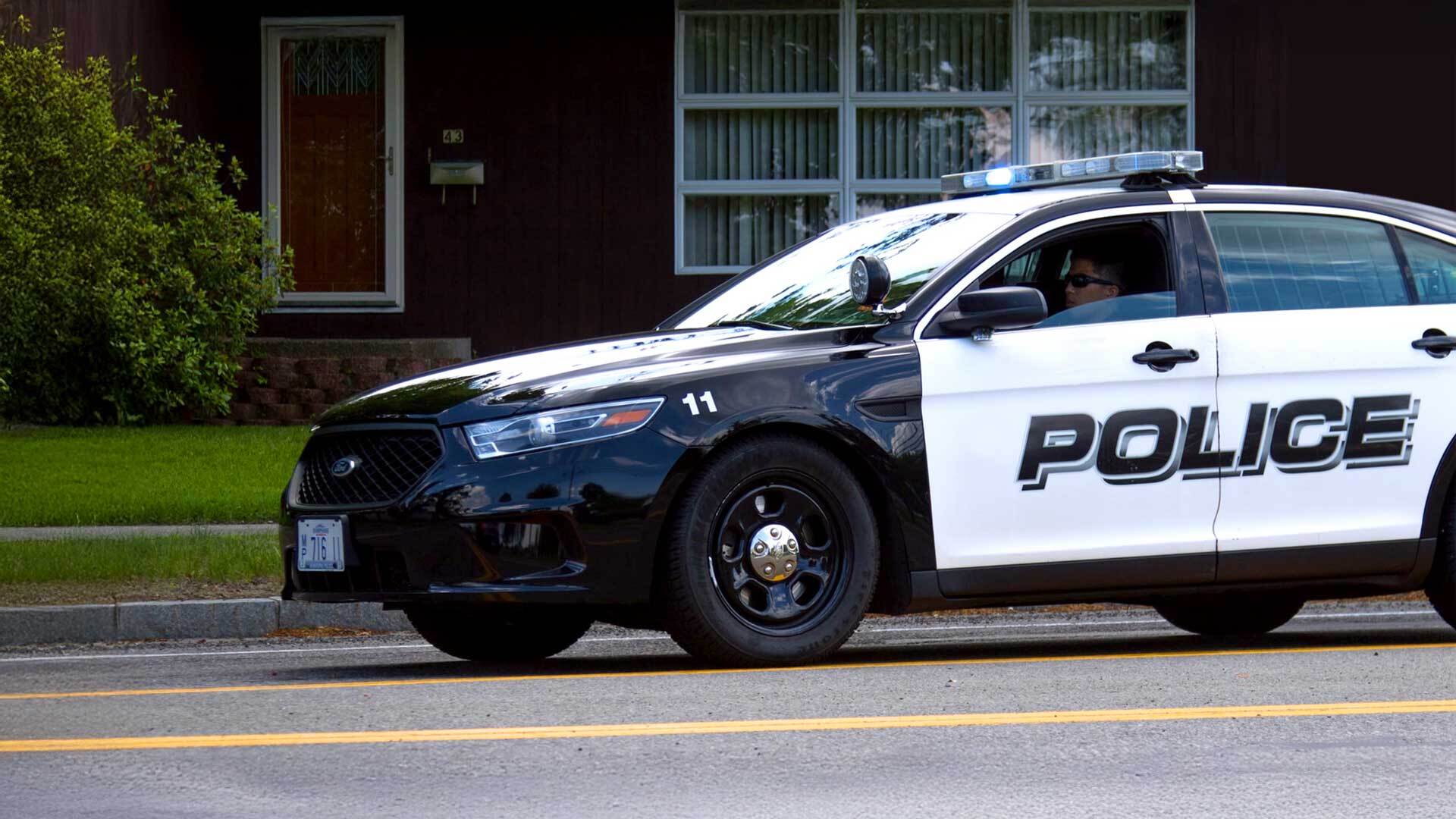 A police officer sits in a black and white police cruiser parked in front of a residential house with a dark wooden door and windows