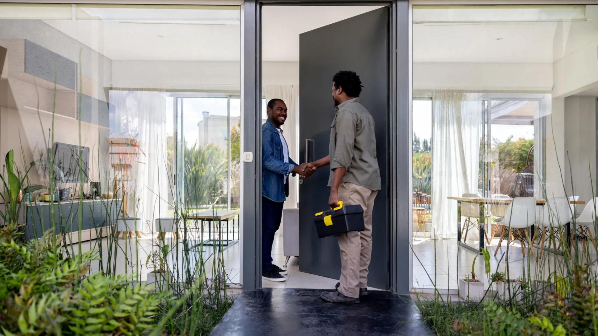 Two men shake hands in a modern entryway with black frame doors, potted plants, and bright windows overlooking a landscape