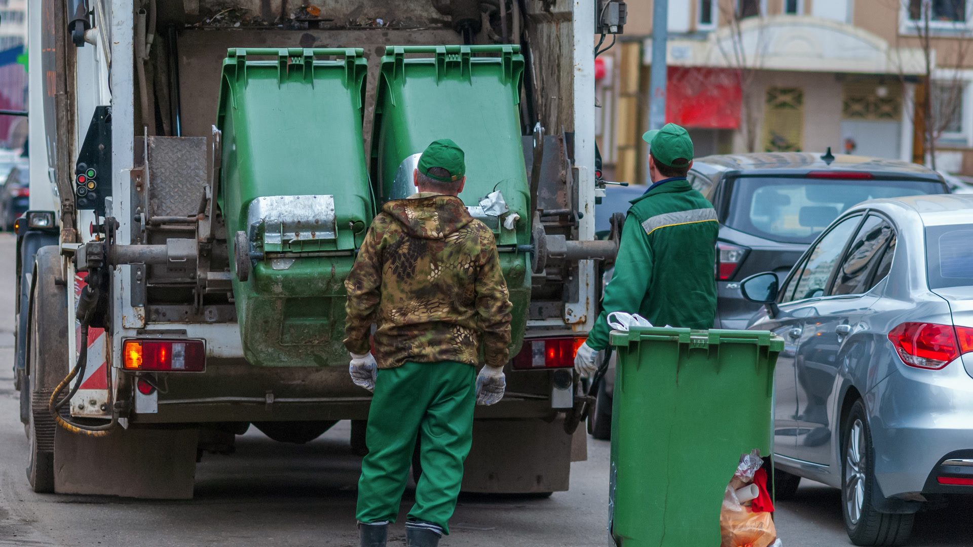 Sanitation worker collecting a residential street-side bin beside a garbage truck.