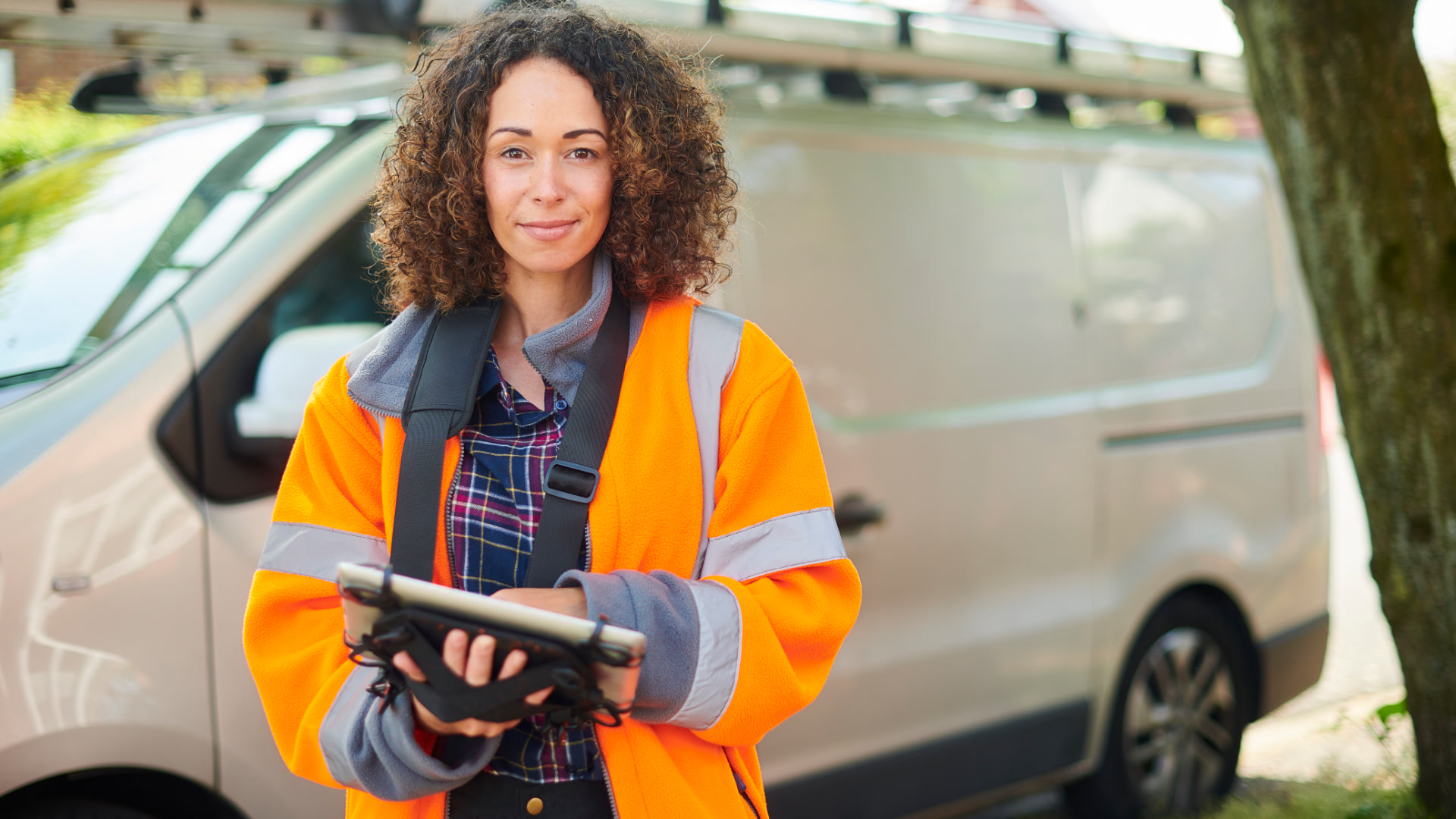 A field service technician in a high-visibility jacket holding a tablet in front of a work van.