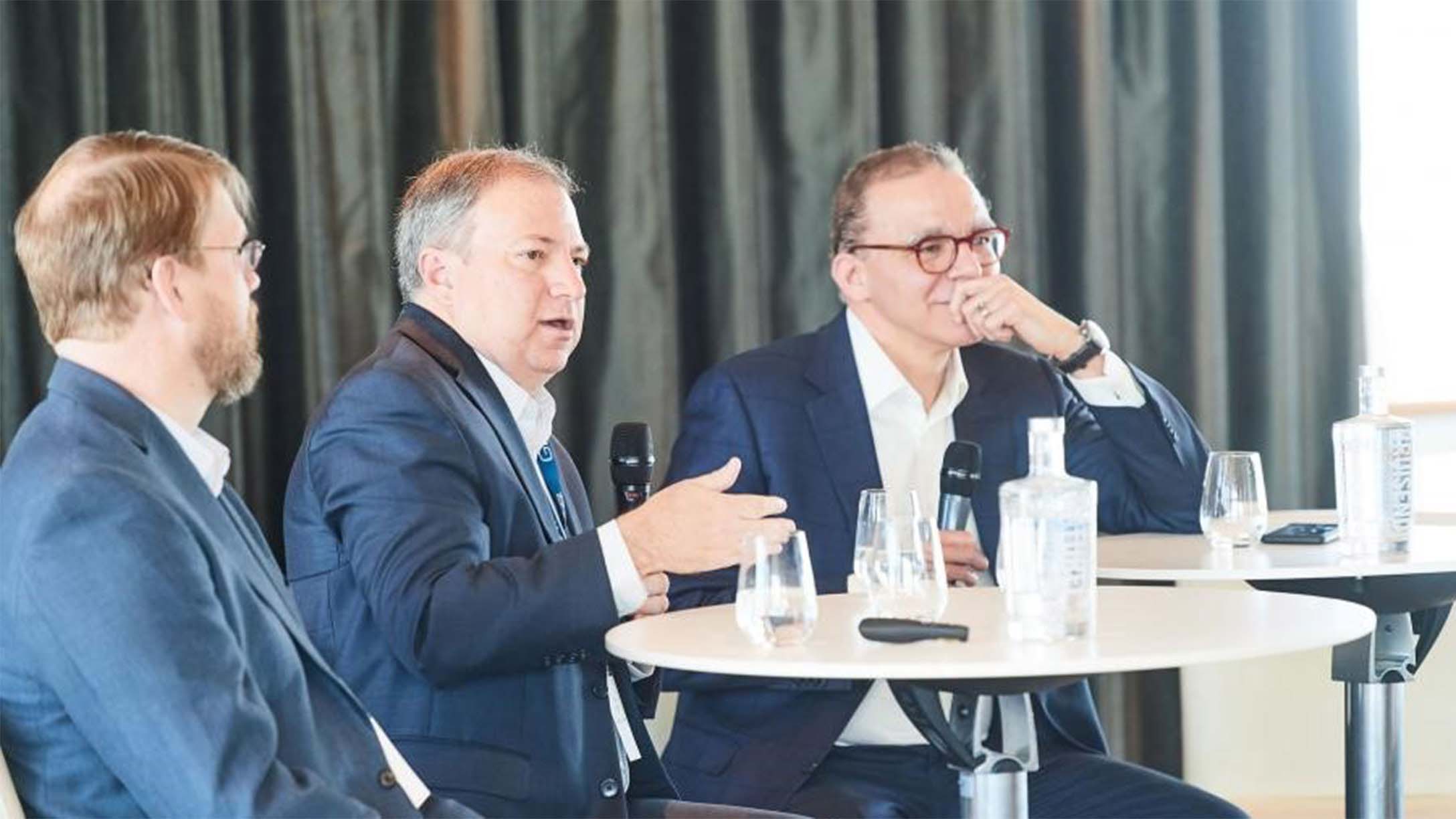 Three men sitting at a table, addressing a room