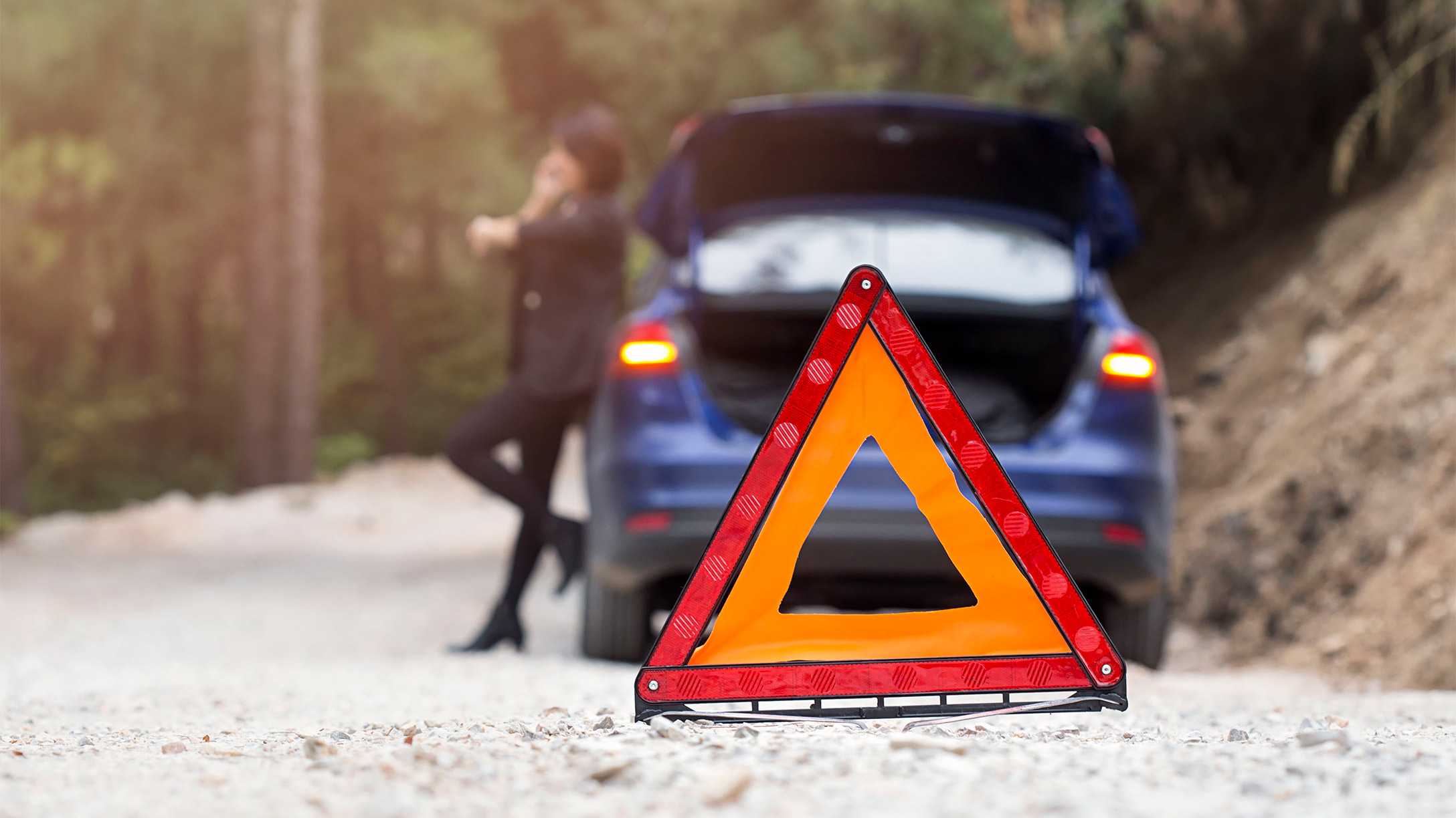 A woman standing against a broken down vehicle with a warning cone in the foreground