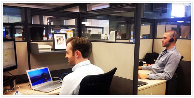 Two male employees sitting at their cubicles, one with a headset on and the other with a laptop open
