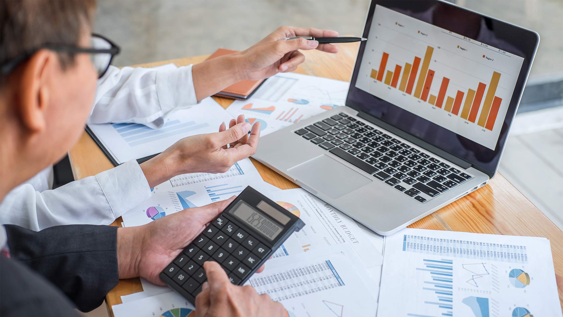 A lady with calculator in her hand is looking at computer with graphs 