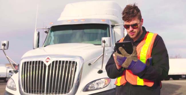 Male driver wearing a high viability vest holding a tablet with a transport truck behind him