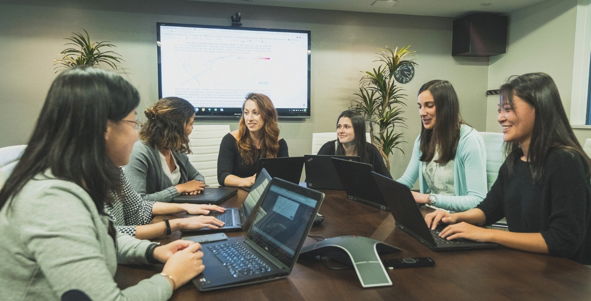 Seven women sitting around a table with laptops open having a discussion