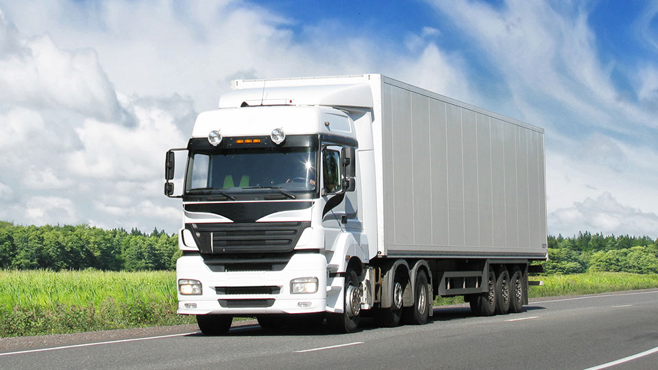 A white coloured transport truck traveling down a road on a sunny day with greenery in the background 