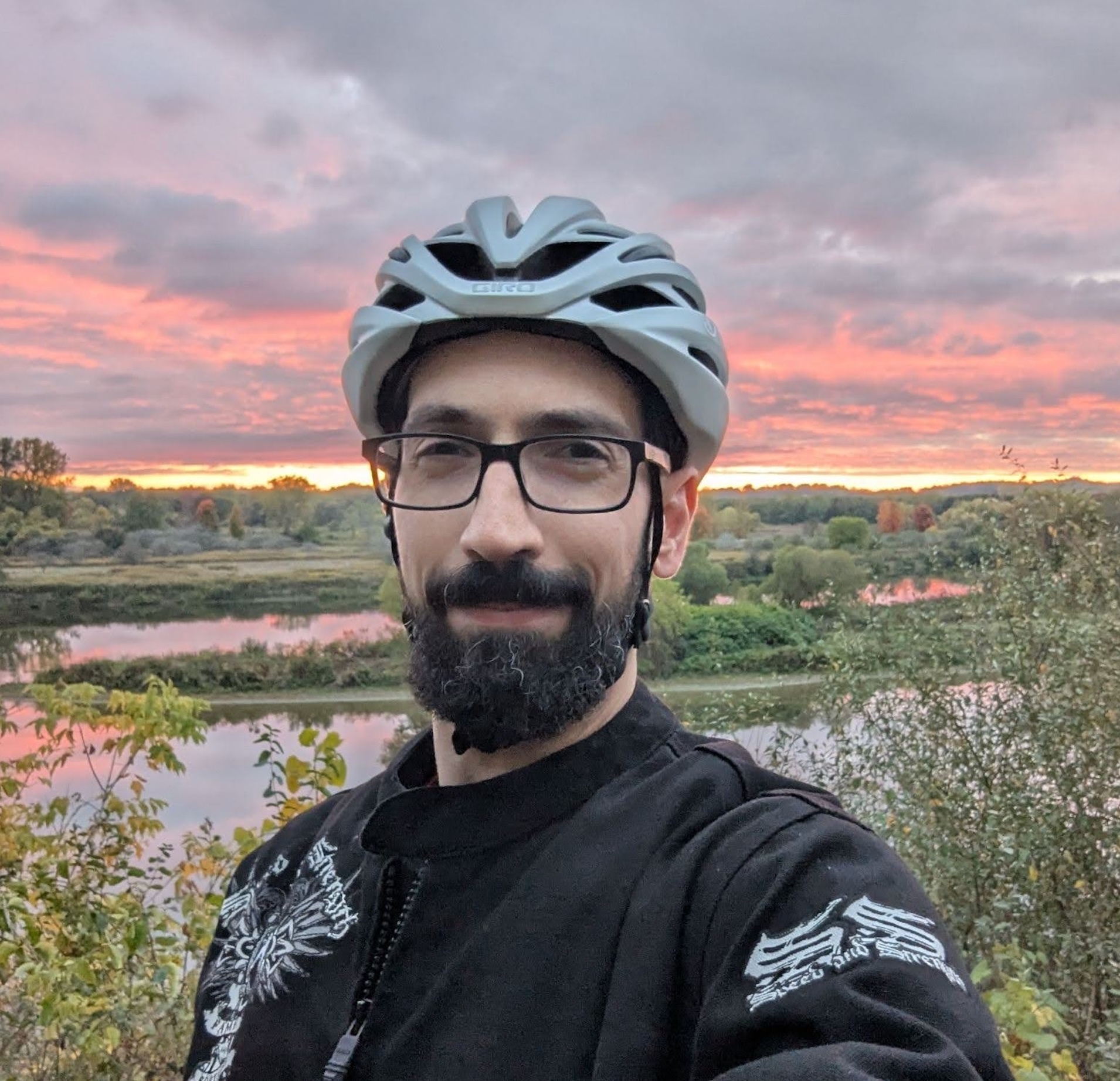 Abdulla Qeblawi smiling outdoors in a bike helmet and glasses, with a river and sunset sky in the background.