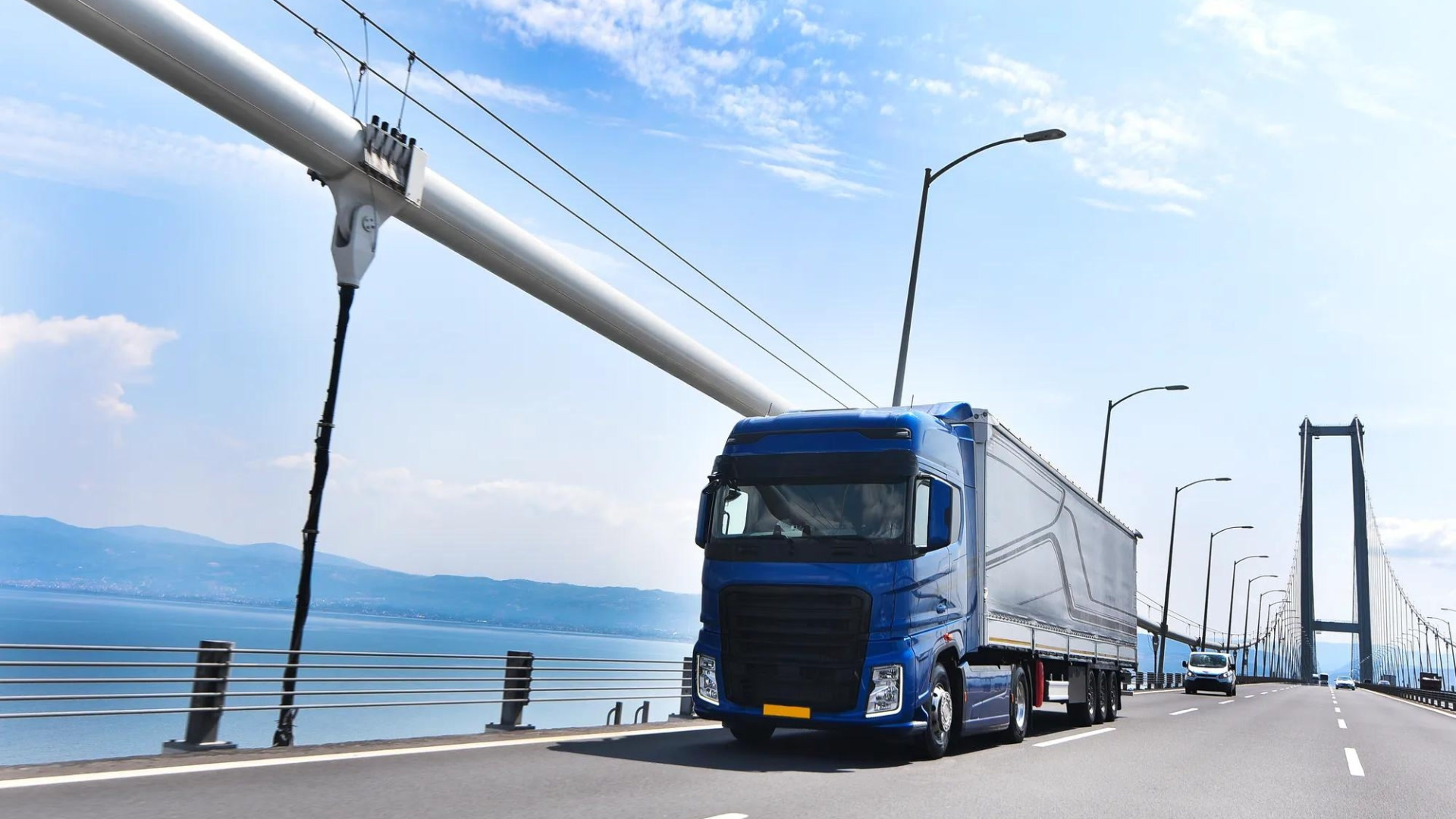 A blue semi-truck travels across a suspension bridge with a scenic coastline and mountains visible in the background