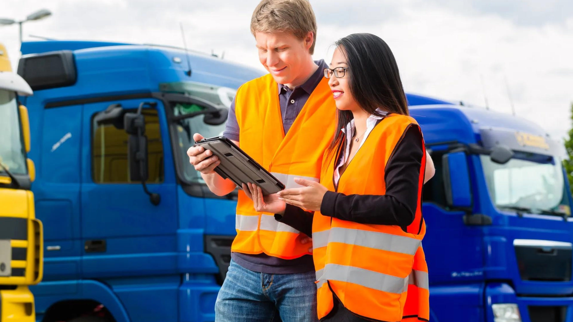 Two workers in orange safety vests review a tablet in front of blue trucks at a logistics facility