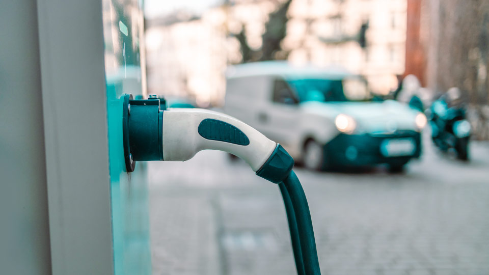 Foto de un enchufe eléctrico azul y blanco conectado a una estación eléctrica con una camioneta blanca en el fondo.