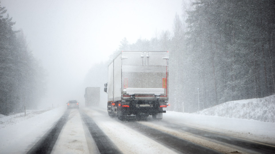 Foto de un camión pesado blanco cubierto en nieve conduciendo por una carretera en un bosque.