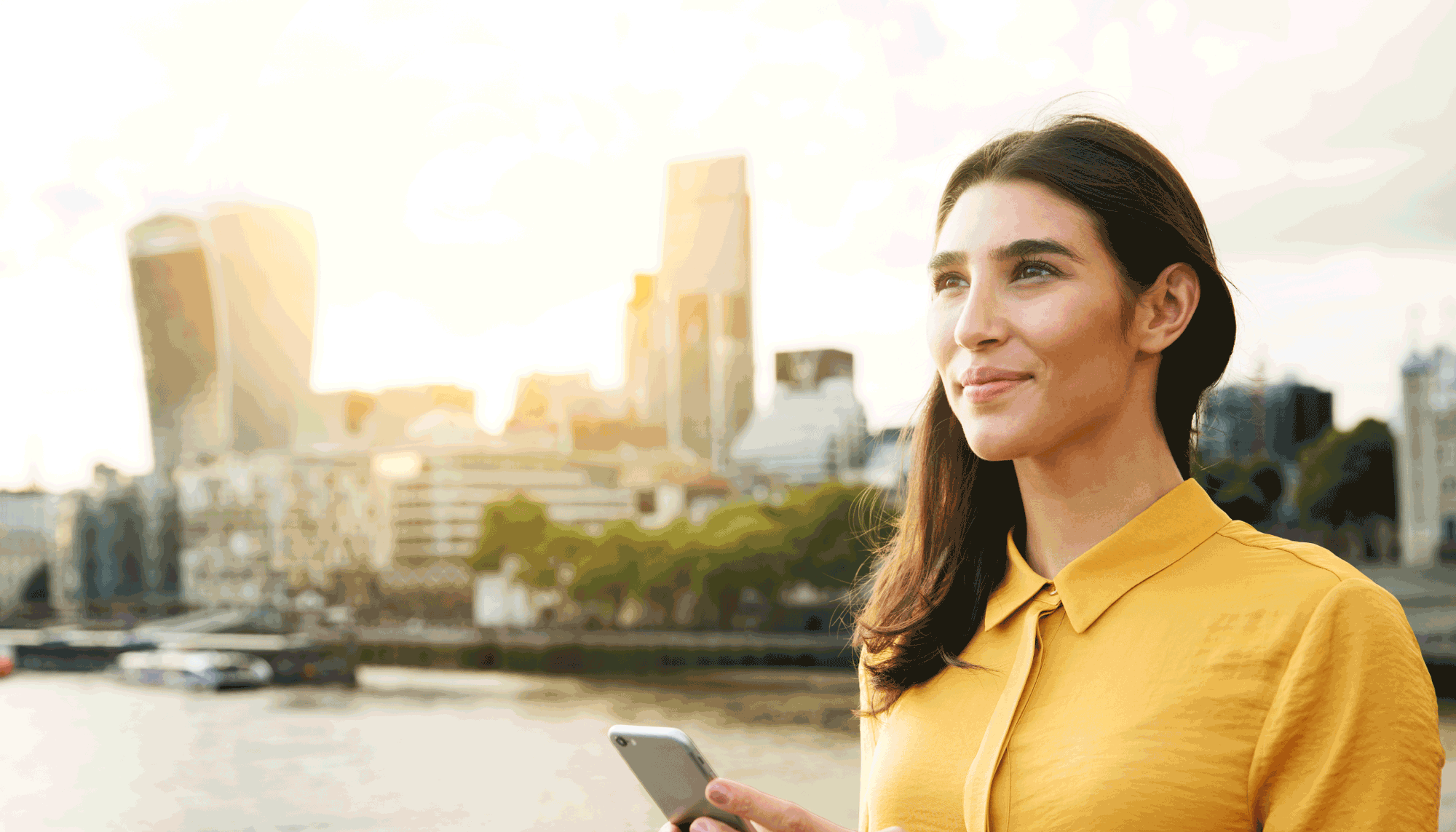 Foto de una mujer con blusa amarilla usando un celular en frente de un lago con una ciudad en el fondo.