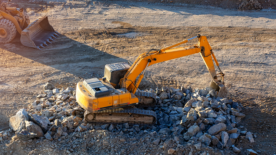 Una excavadora amarilla de orugas está trabajando en una obra de construcción, moviendo y apilando grandes rocas. Al fondo se ve parte de otra máquina pesada.