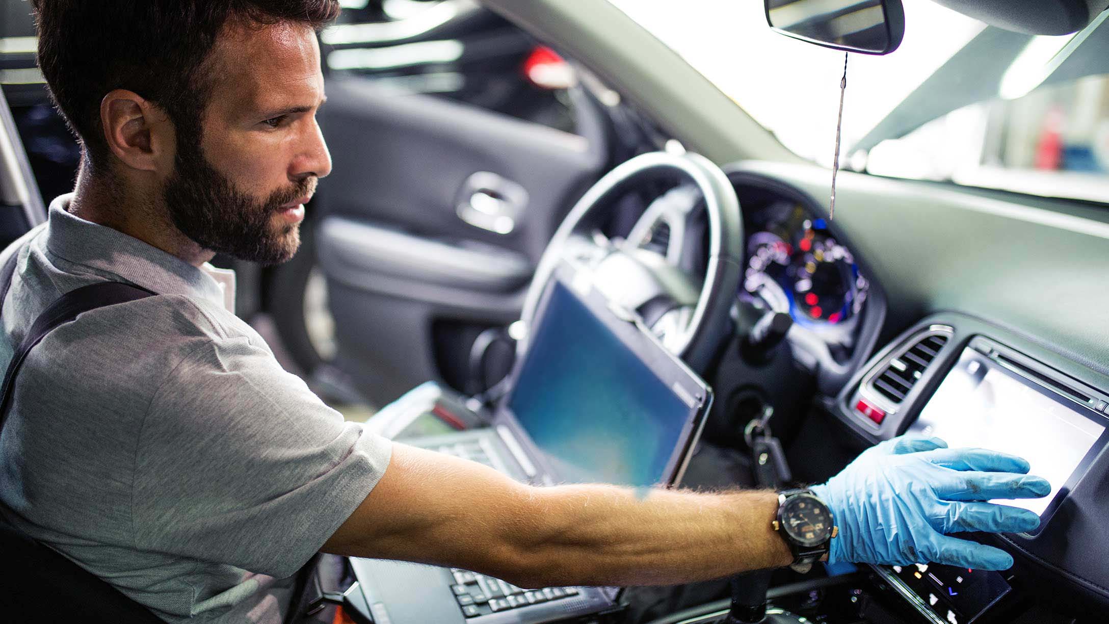 un hombre en el asiento del conductor de un vehículo, concentrado en una laptop que tiene abierta frente a él. Parece estar realizando algún tipo de diagnóstico o configuración, ya que también extiende una mano enguantada 
