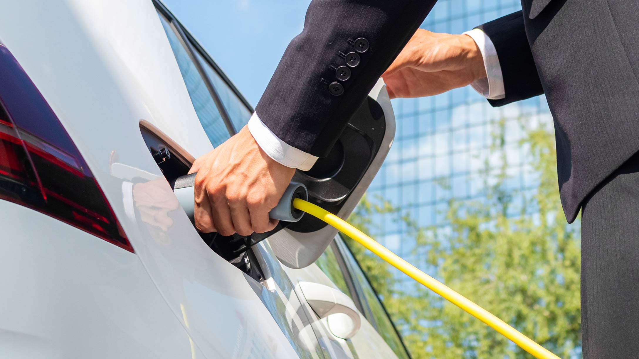 Imagen de un hombre con traje conectando el cargador de un vehículo eléctrico a su automóvil blanco en un día soleado con un cielo azul de fondo.