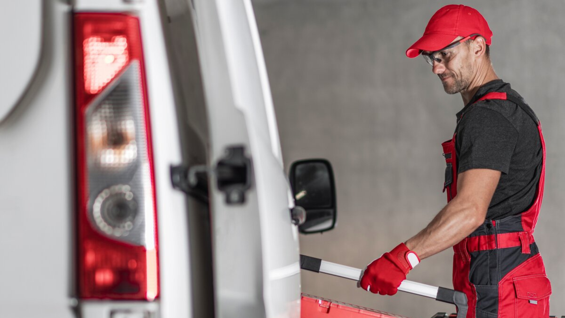 Un technicien en salopette rouge prend une boîte à outils rouge et noire à l'arrière ouvert d'une fourgonnette de service blanche.