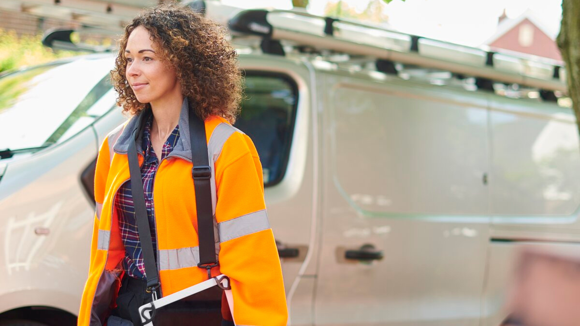 Una trabajadora de la construcción con un casco y un chaleco de seguridad está de pie frente a una furgoneta de trabajo, sosteniendo una tableta y una caja de herramientas.