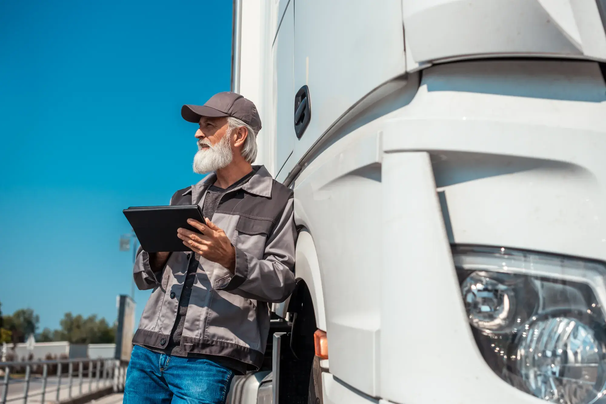 Chauffeur de camion senior, appuyé contre son camion blanc tout en tenant une tablette.