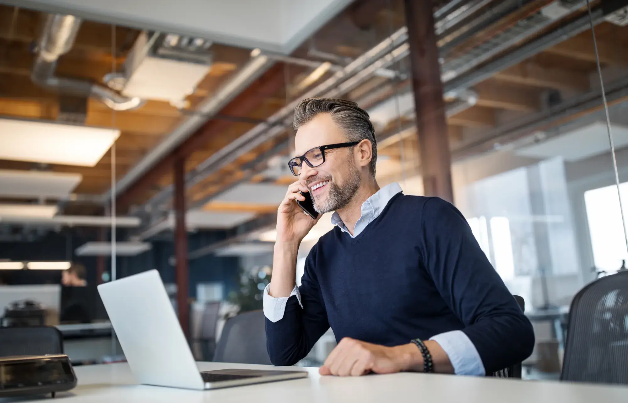 Homme d'affaires souriant portant des lunettes, au téléphone devant un ordinateur portable dans un bureau moderne.