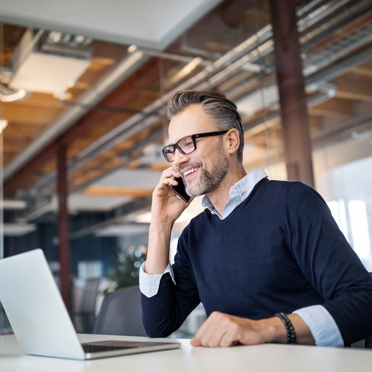 Homme d'affaires souriant portant des lunettes, au téléphone devant un ordinateur portable dans un bureau moderne.