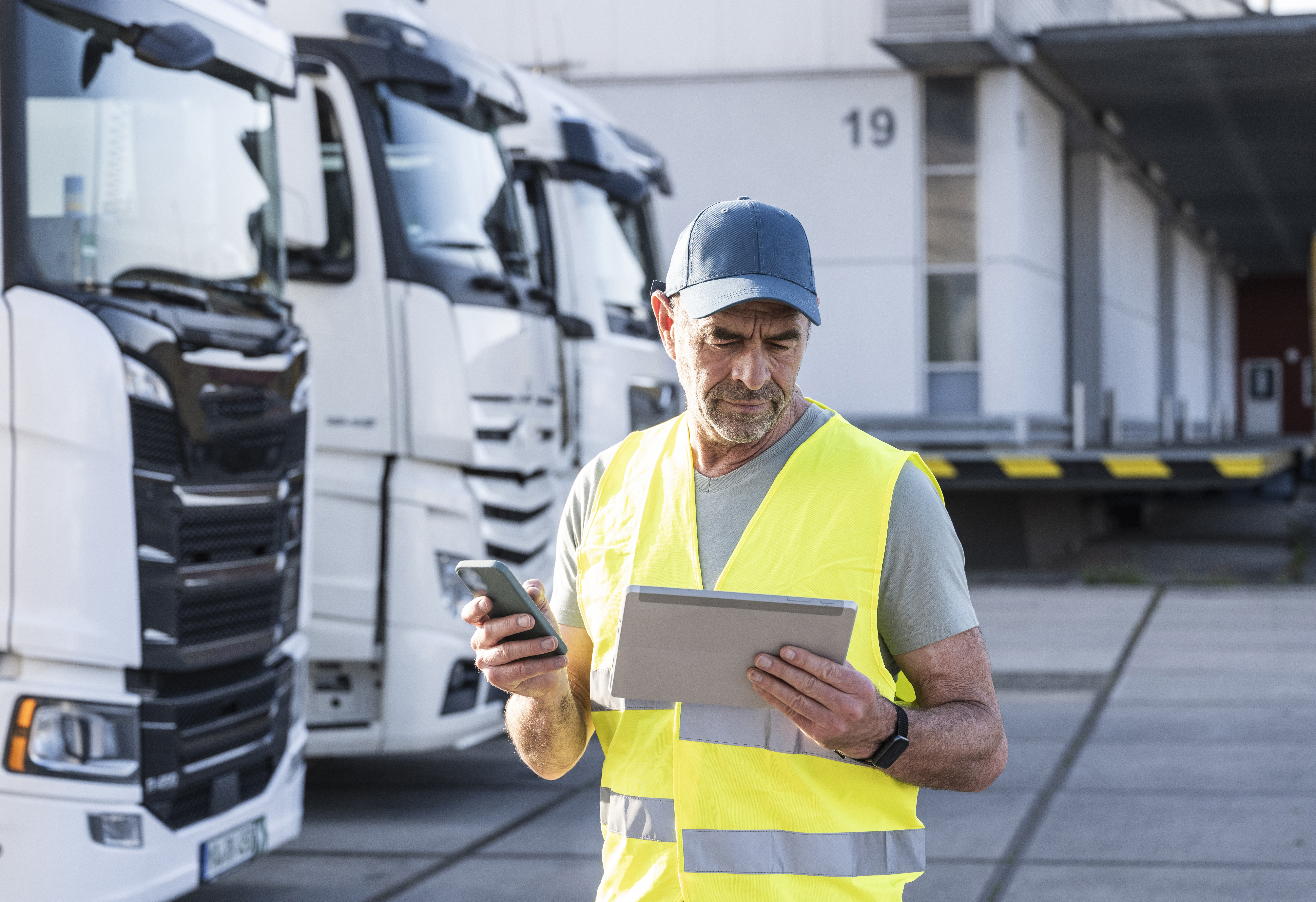 Conducteur portant un gilet jaune et une casquette qui regarde une tablette et tient un portable, devant des camions blancs.