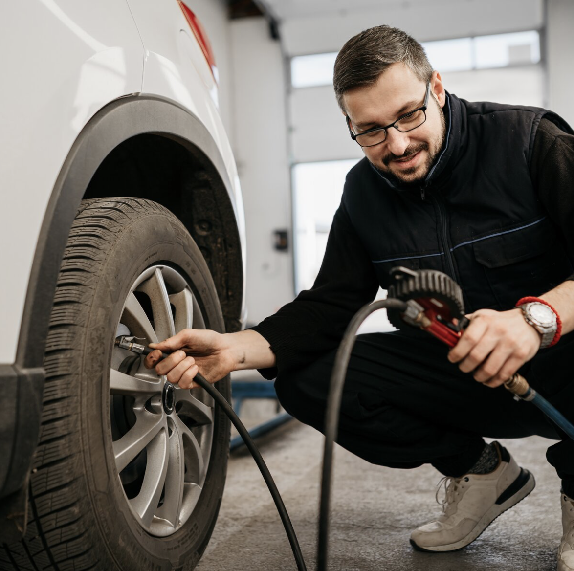 Un mécanicien, portant des lunettes et une tenue de travail, est accroupi pour vérifier et gonfler le pneu d'une voiture blanche.