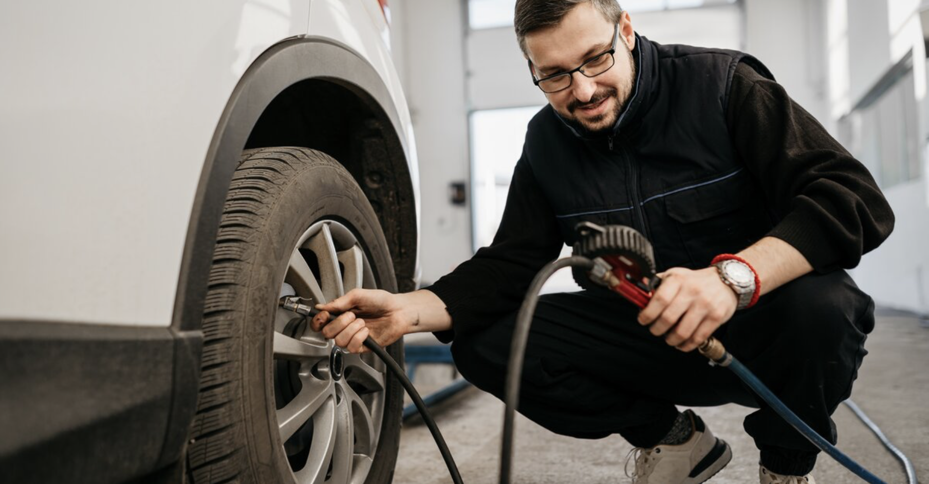 Un mécanicien, portant des lunettes et une tenue de travail, est accroupi pour vérifier et gonfler le pneu d'une voiture blanche.
