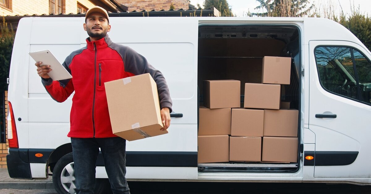 Un livreur en veste rouge et casquette se tient à l'extérieur d'une camionnette de livraison blanche avec la porte arrière ouverte. Il tient une tablette d'une main et une boîte en carton de l'autre. Des piles de colis sont visibles dans la camionnette.