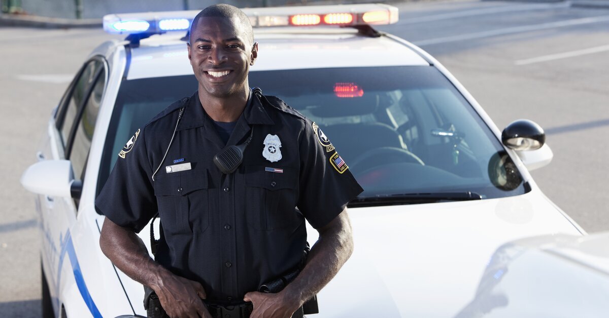 Policier souriant devant une voiture de police blanche.