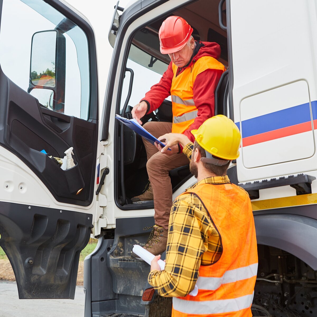 Deux chauffeurs de camion en gilets de sécurité et casques (un assis dans la cabine du camion, l'autre debout à l'extérieur) discutent une liste de contrôle.