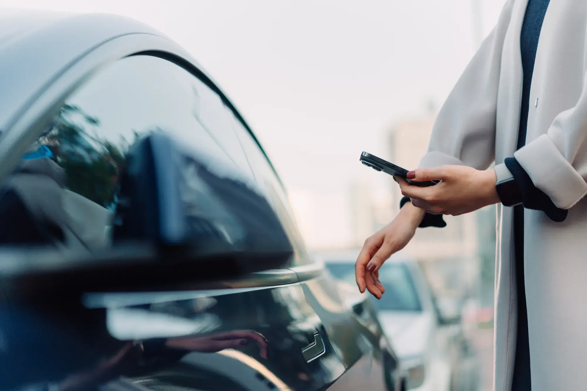 Gros plan sur les mains d'une femme portant un manteau qui tient son portable face à une voiture foncée.