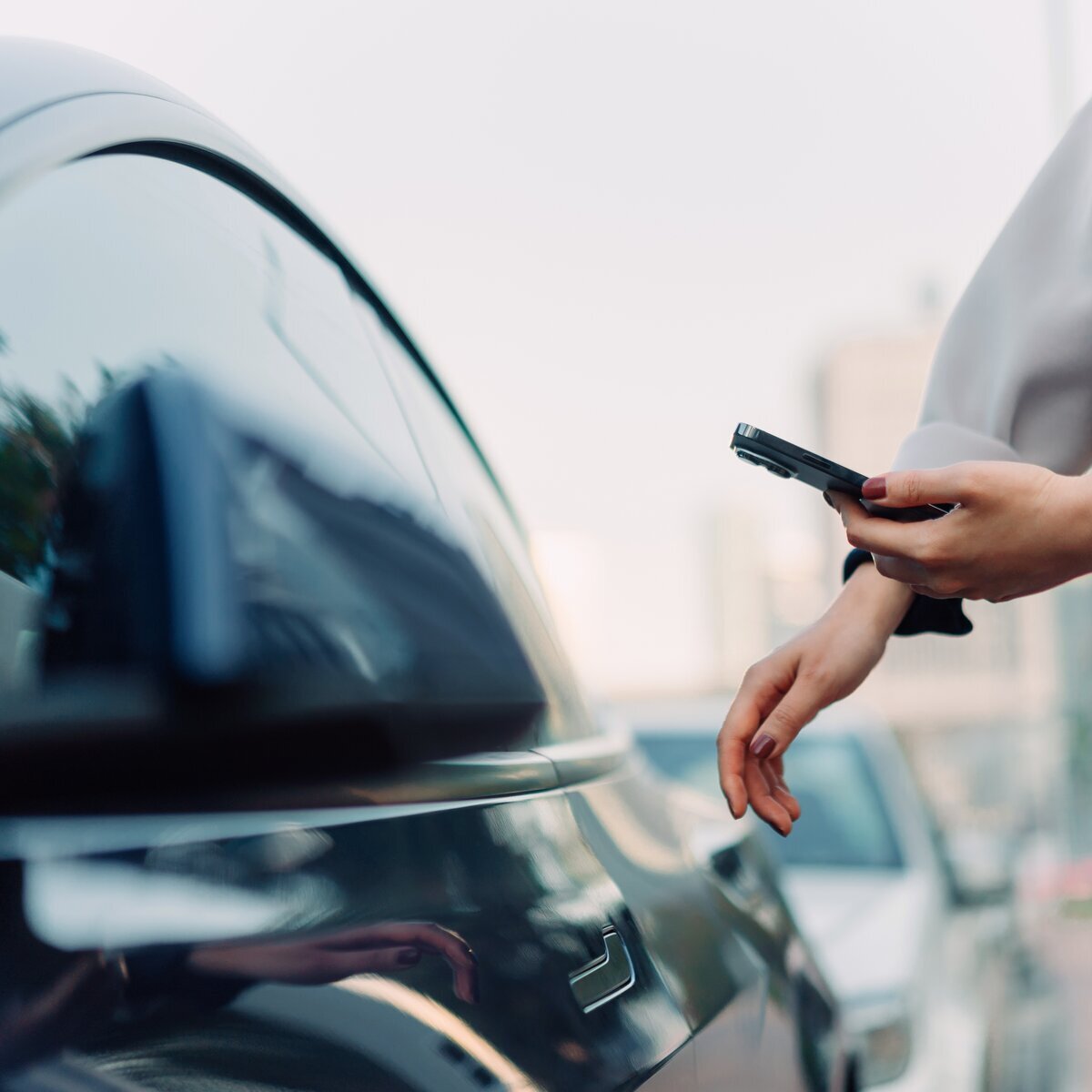 Gros plan sur les mains d'une femme portant un manteau qui tient son portable face à une voiture foncée.