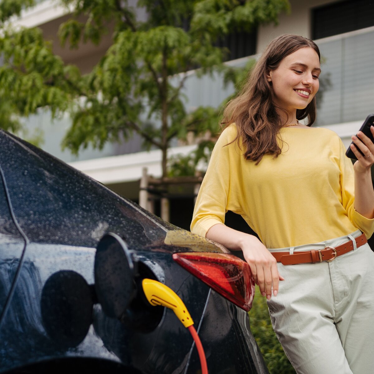 Femme souriante utilisant son portable devant un véhicule électrique qui charge.