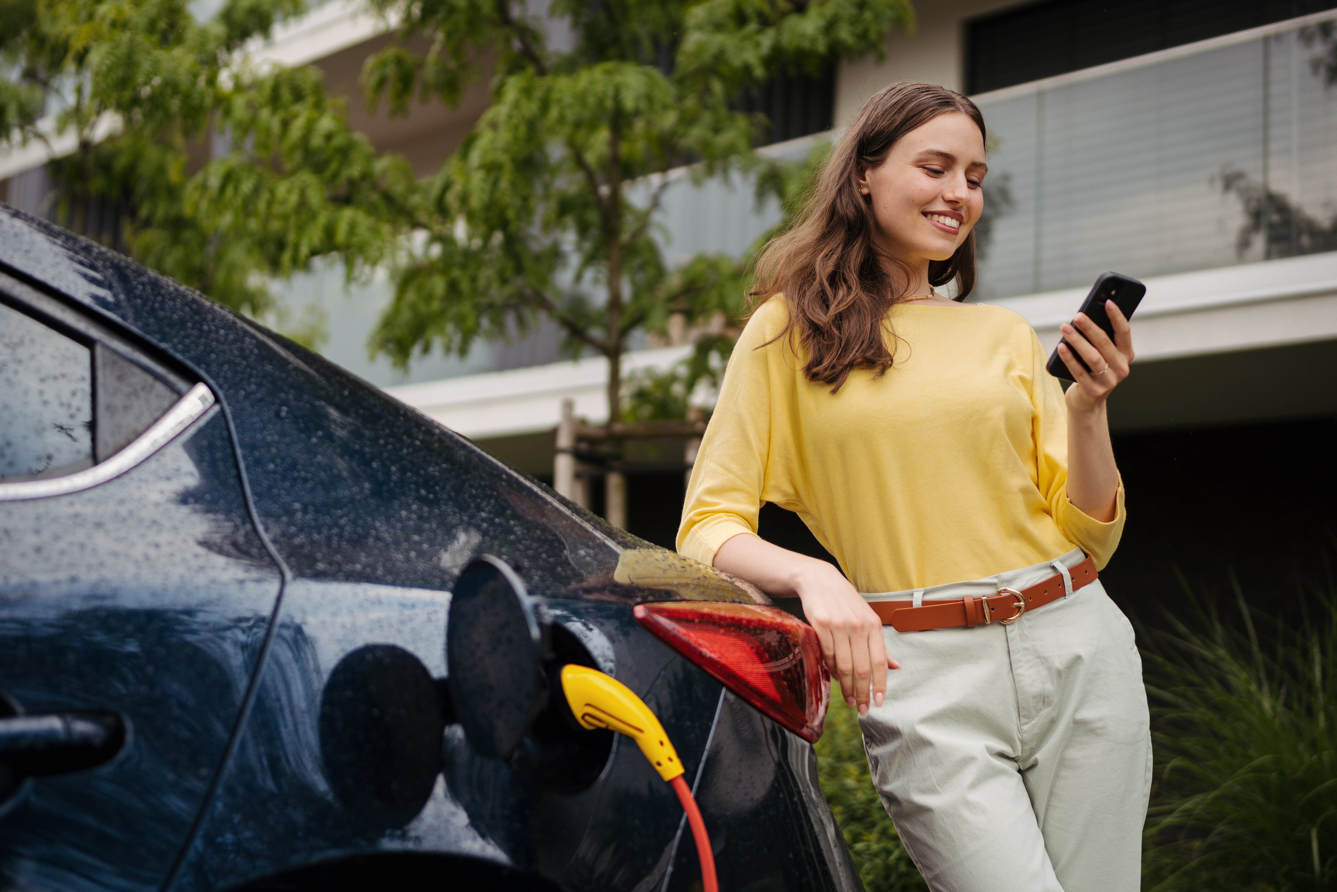 Femme souriante utilisant son portable devant un véhicule électrique qui charge.