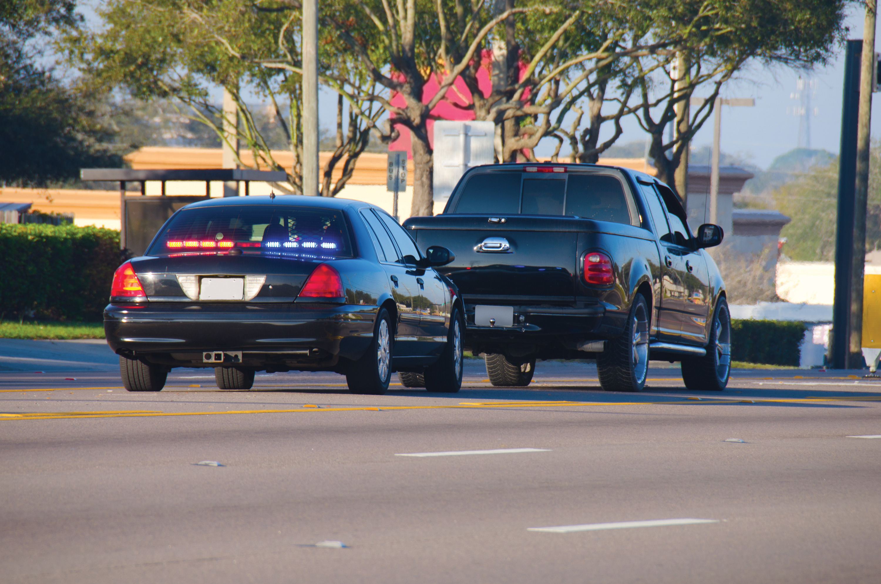 Une voiture de police noire avec des gyrophares allumés, suivie de près par un pick-up noir, roulant sur une route.